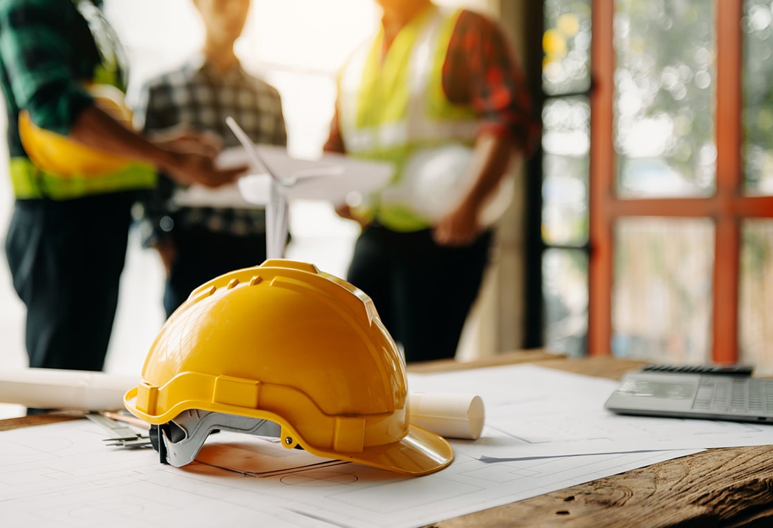A yellow hard hat is placed on a wooden table. There are blueprints and a laptop in the background. Two people wearing reflective vests and one person holding a drone are visible in the background. The blueprints are rolled up and the laptop is open. 