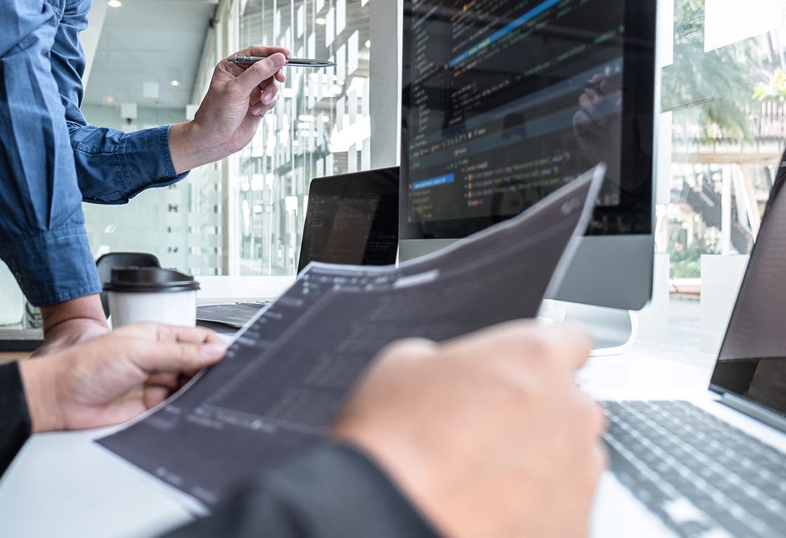 Two people working in a modern office setting. The person on the left is holding a pen and pointing at a computer screen. The person on the right is holding a document and looking at it. 