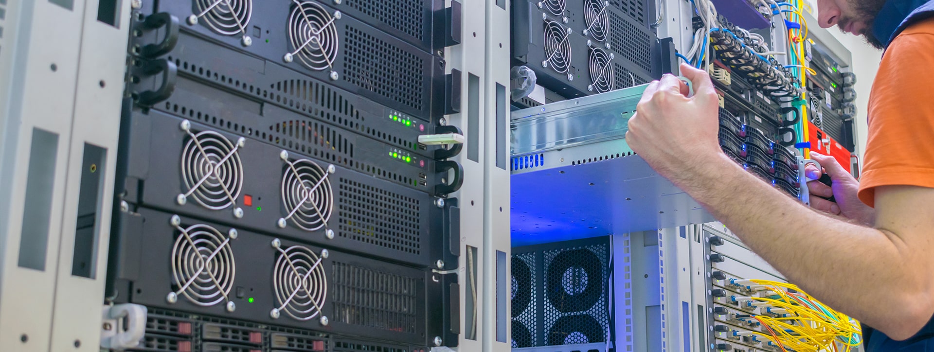 A person working on a server rack in a data center. The server rack is black and has multiple rows of fans on the back. The person is wearing an orange shirt and is holding a blue tool. The person is working on a server that is partially pulled out of the rack. There are multiple cables visible in the background.