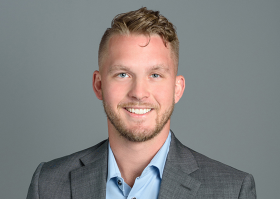 A man, Jared Hill, is wearing a gray suit jacket and a blue collared shirt.